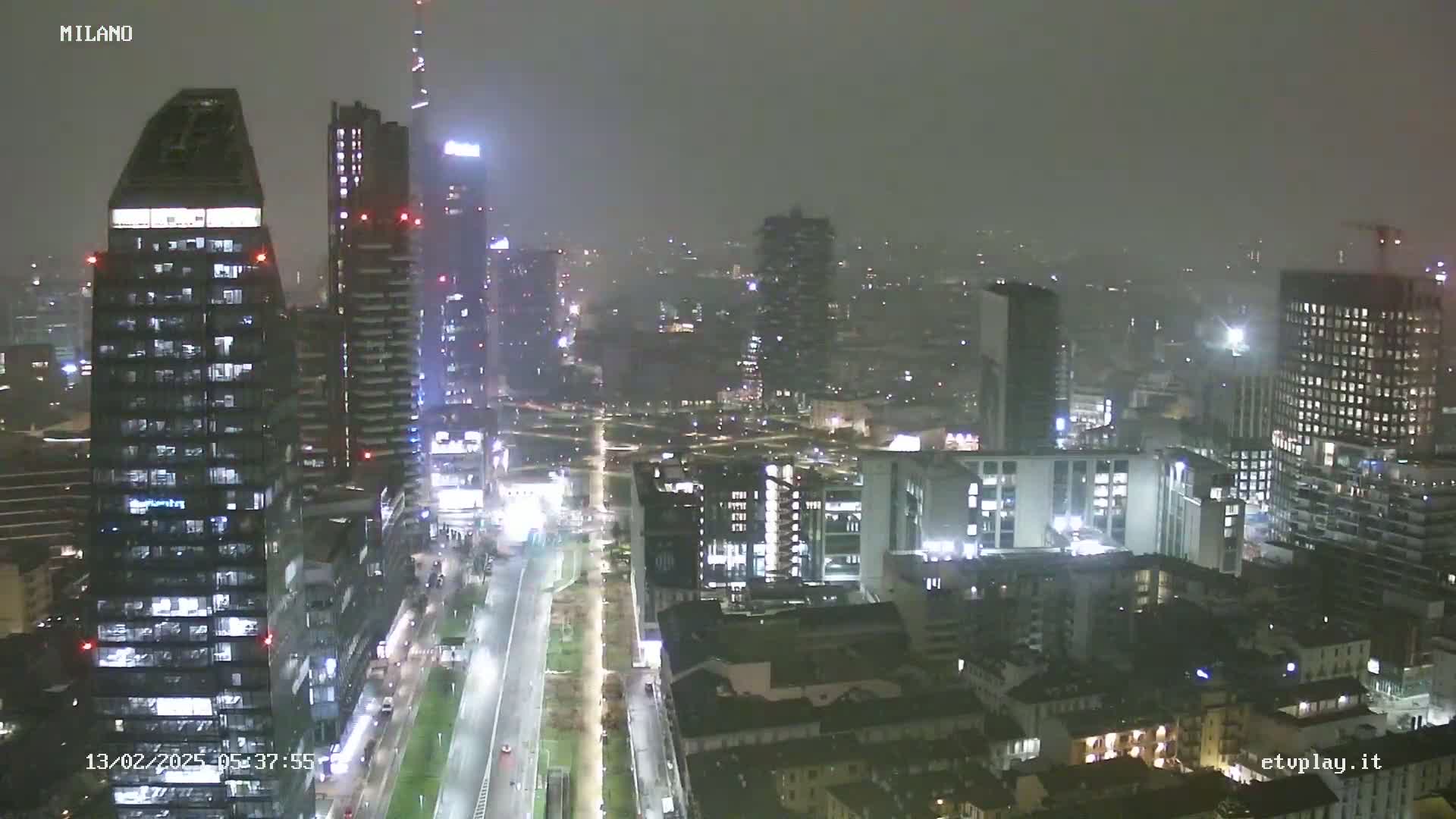 A nighttime, hazy view of a city skyline, showing numerous skyscrapers and buildings illuminated against a dark, overcast sky.