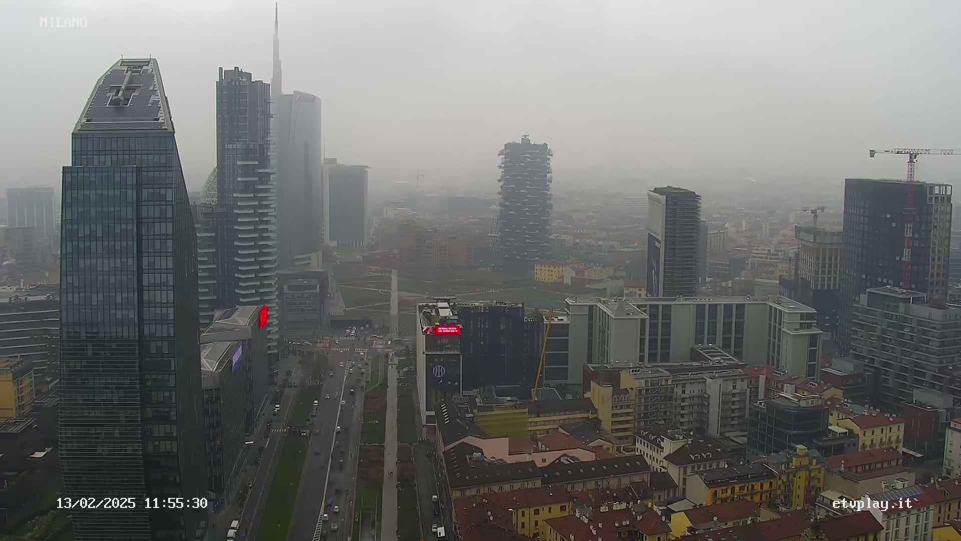A hazy, overcast cityscape view shows numerous tall modern buildings and shorter, older buildings, with a street and some cars visible below.