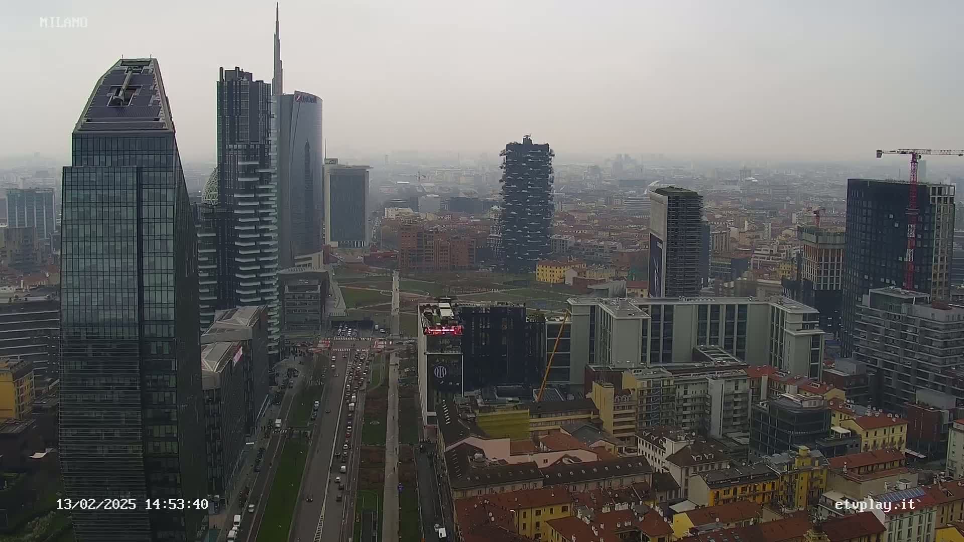 A hazy cityscape view of Milan shows numerous modern skyscrapers and high-rise buildings, with several under construction, along with a street with moving vehicles below.
