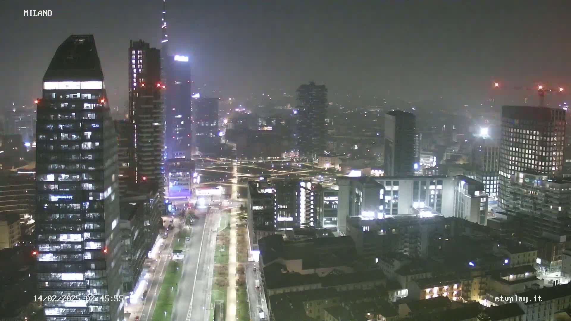 A nighttime aerial view of a city skyline with hazy weather conditions, showing numerous illuminated skyscrapers and buildings.