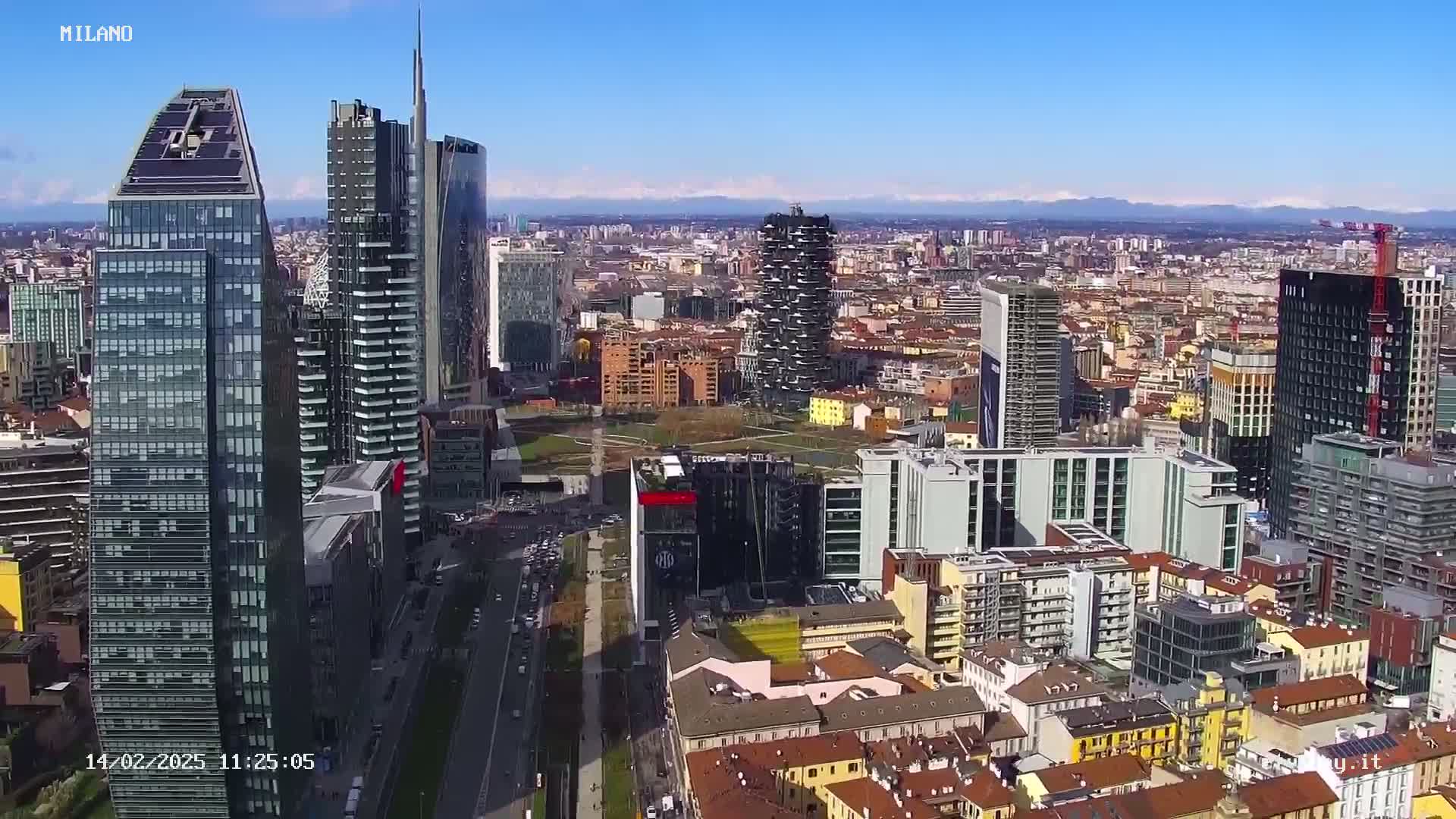 An aerial view of a city with numerous skyscrapers under a clear blue sky, mountains visible in the distance.