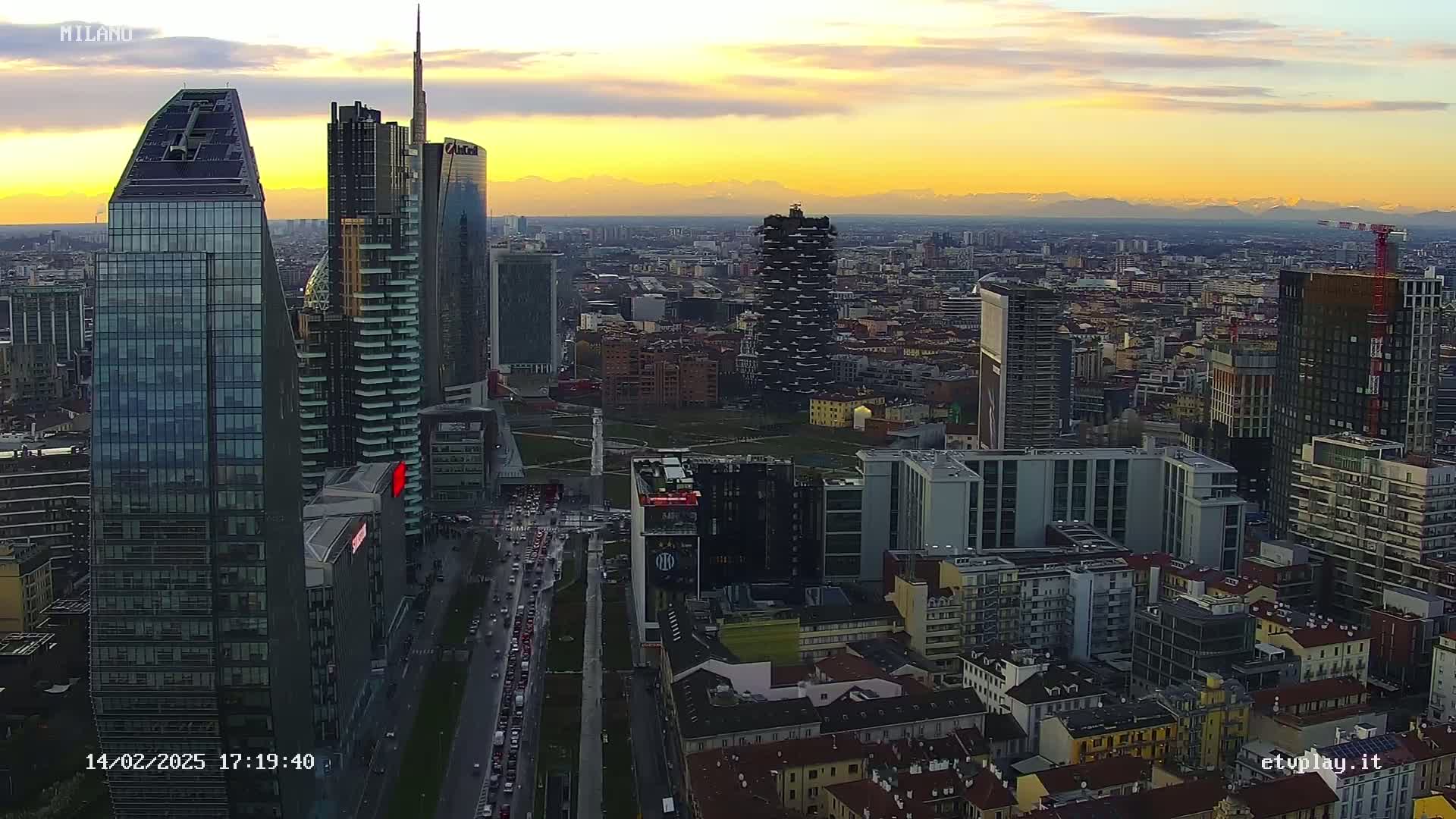 An aerial view of a dense cityscape at sunset, with mountains visible in the distance under a partly cloudy sky.