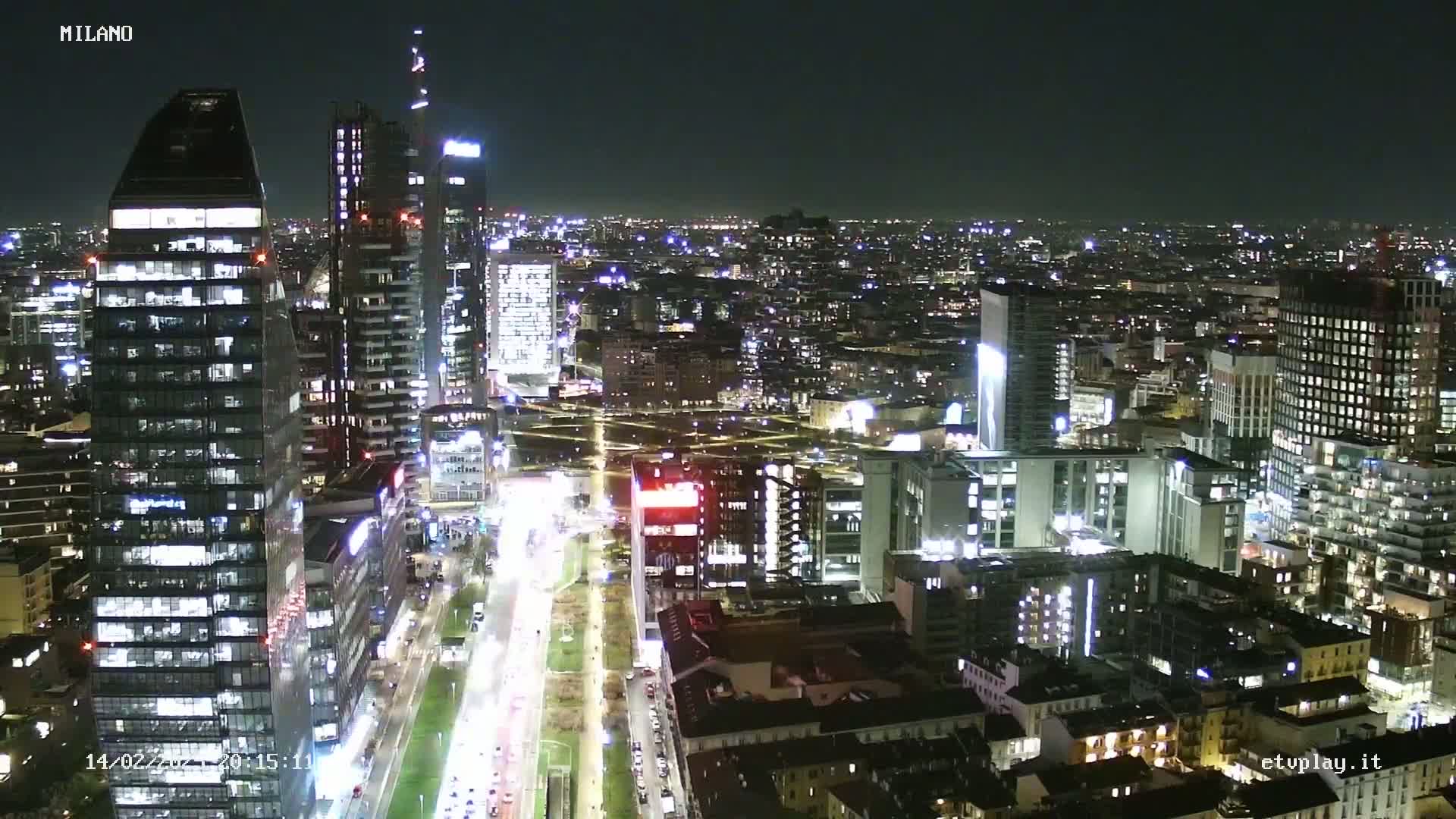 A nighttime aerial view of a city, showing brightly lit skyscrapers and streets with moving traffic under a clear, dark sky.