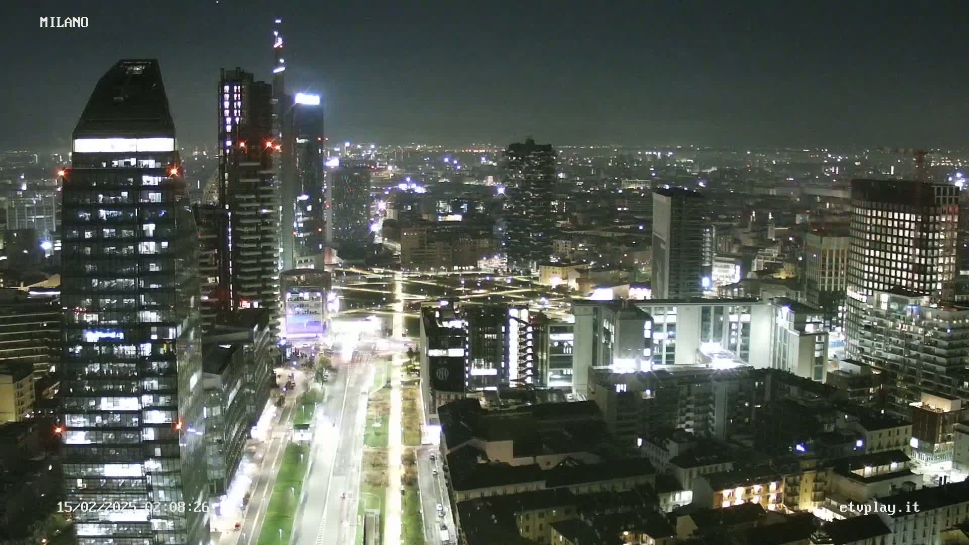 A nighttime aerial view of a city's skyline, showing illuminated skyscrapers and streets with moving vehicles under a clear sky.
