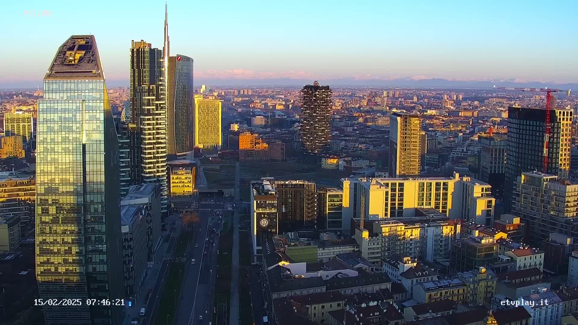 An aerial view of a cityscape at sunset, showing numerous skyscrapers and high-rise buildings, with mountains visible in the distance under a clear sky.
