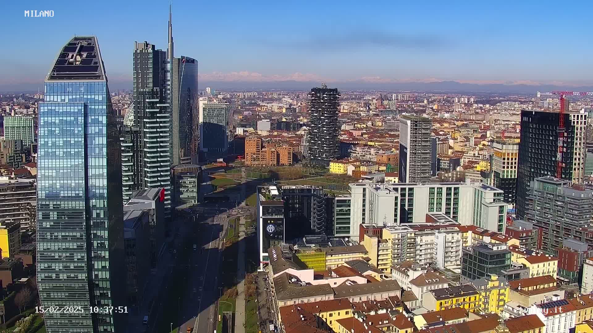 A high-angle, sunny-day view of a dense cityscape featuring numerous skyscrapers and high-rise buildings, with distant mountains visible on the horizon.