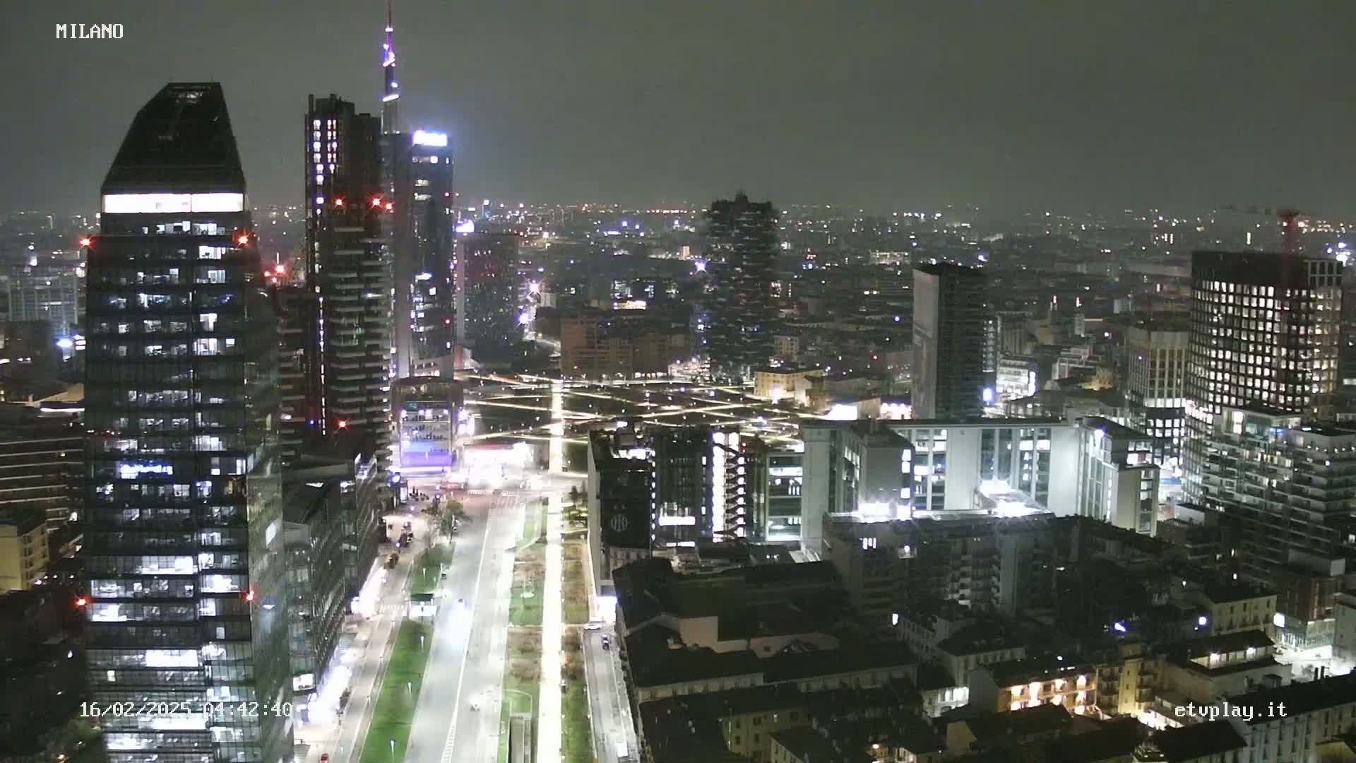 A nighttime aerial view of a city skyline with many illuminated skyscrapers and buildings, under a dark, overcast sky.