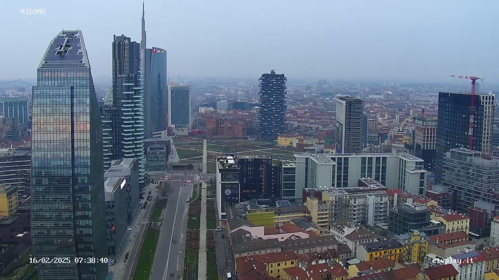 An aerial view of a dense cityscape under a hazy sky shows numerous skyscrapers and buildings of varying heights and architectural styles.