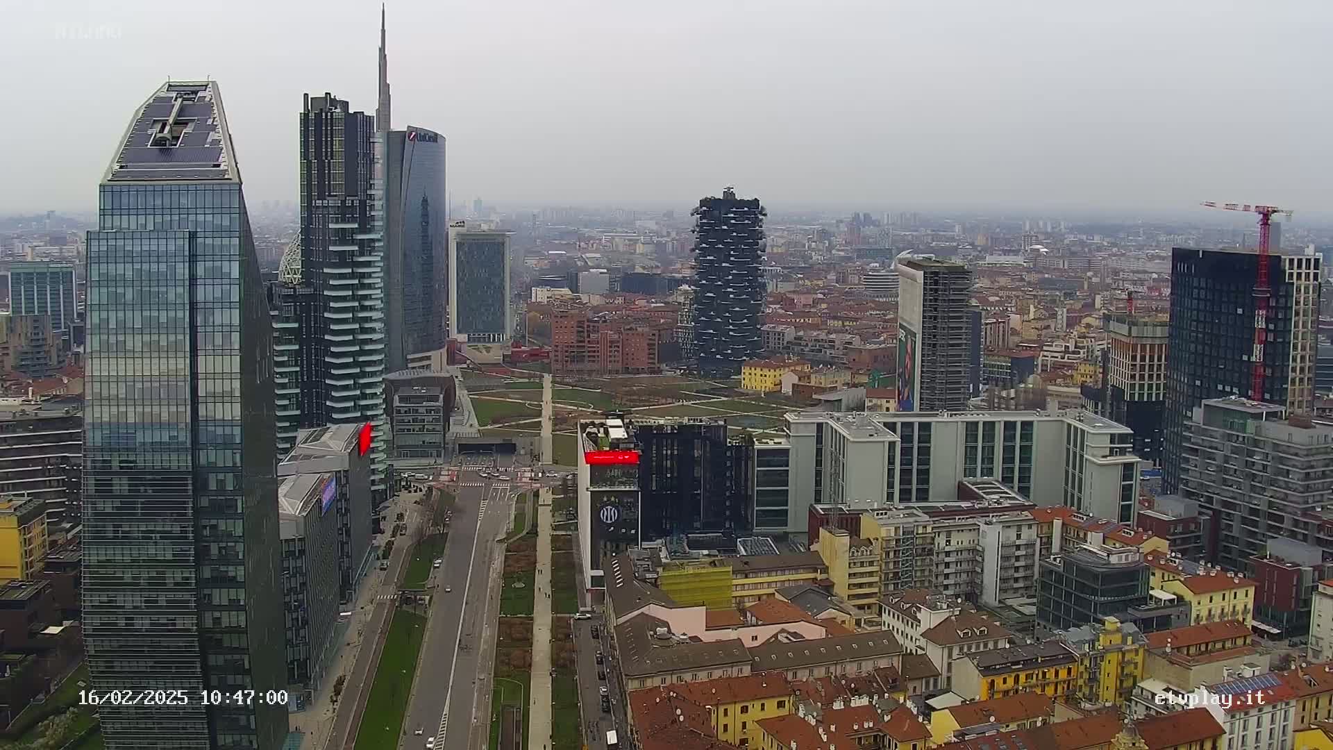 A high-angle, long shot of a cityscape on an overcast day shows many modern skyscrapers and traditional buildings, with a large road running through the center.