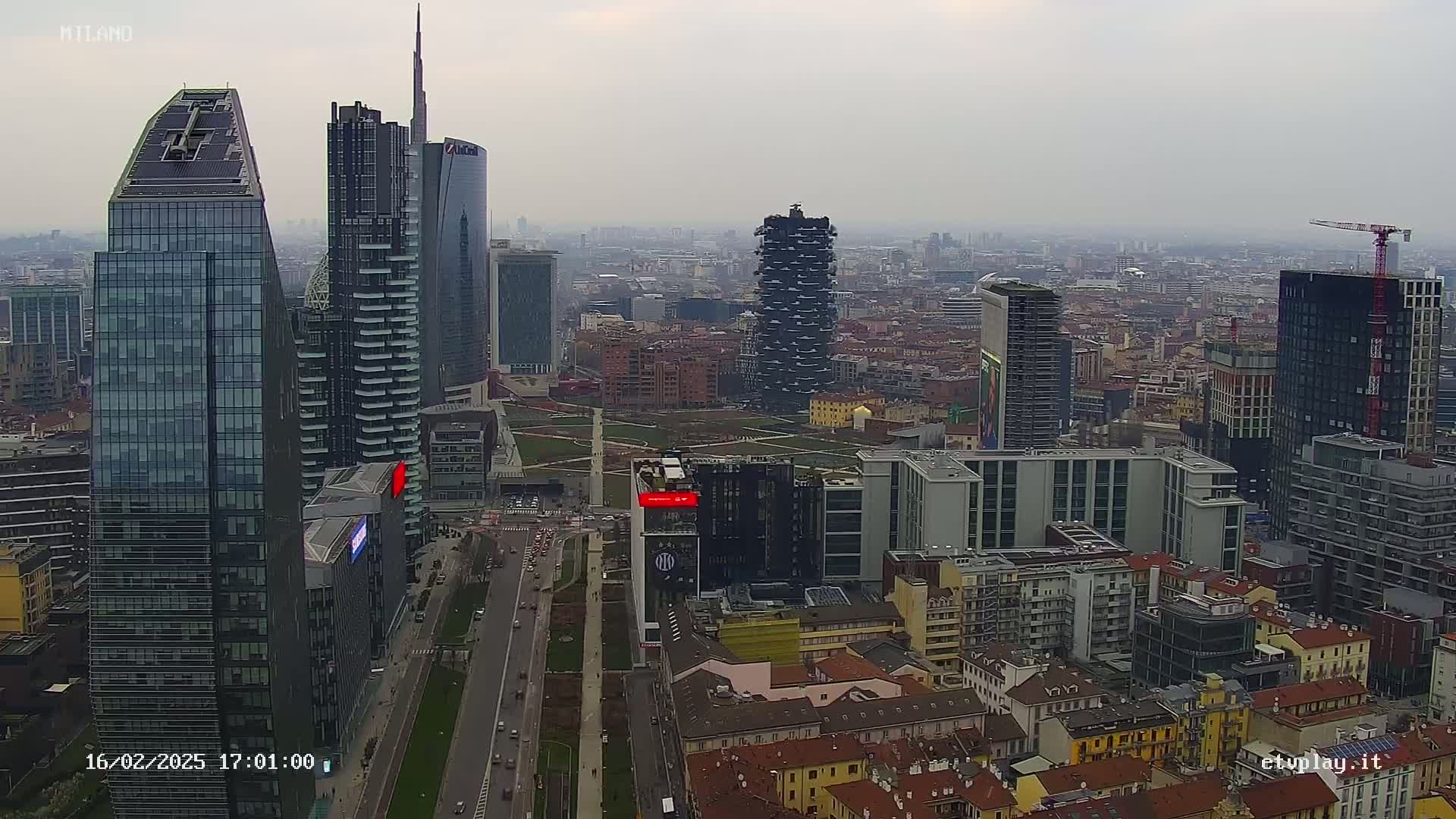 A high-angle, long shot of a cityscape on an overcast day, showing numerous skyscrapers and surrounding buildings, with a wide road running through the center.