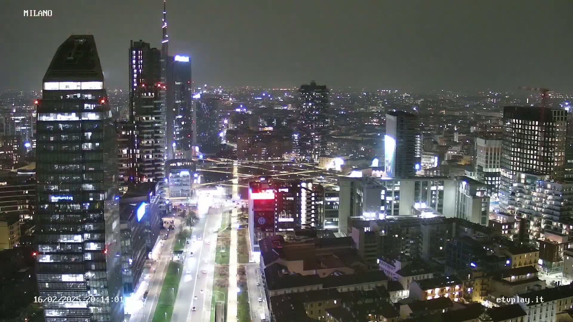 A nighttime aerial view of a city with many illuminated skyscrapers and streets, under a dark, cloudy sky.