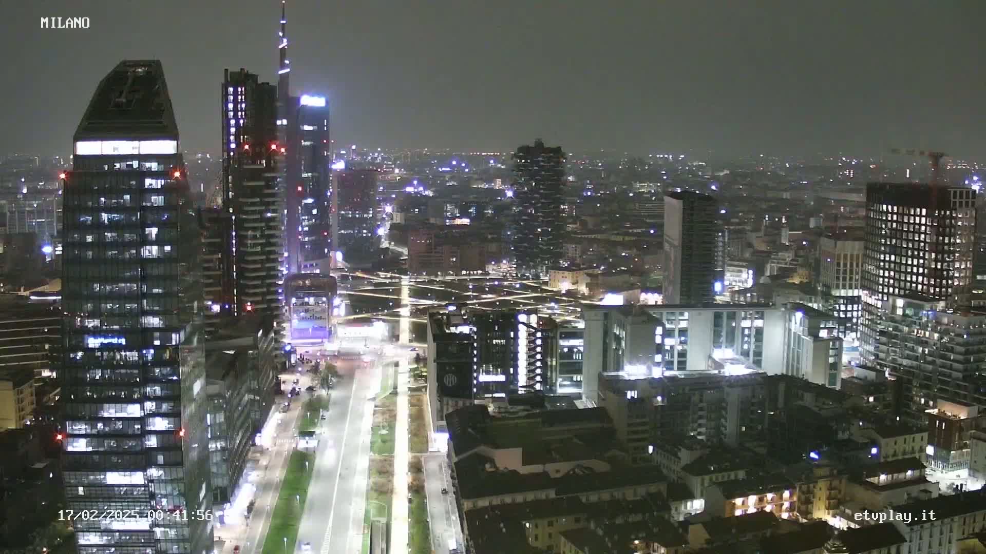 A nighttime aerial view of a city, showing numerous illuminated skyscrapers and buildings, with a mostly clear but dark sky.