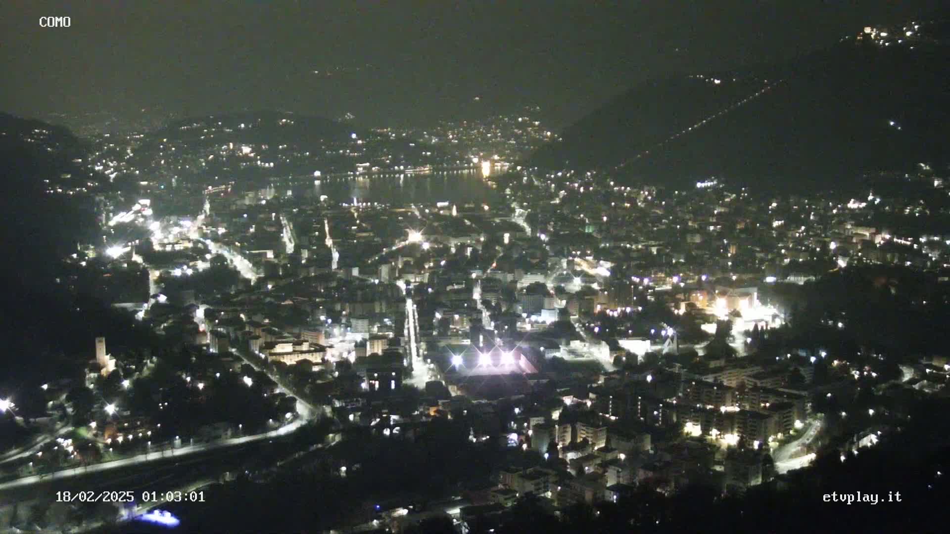 A nighttime, high-angle view of a city nestled in a valley next to a body of water, illuminated by streetlights and building lights.