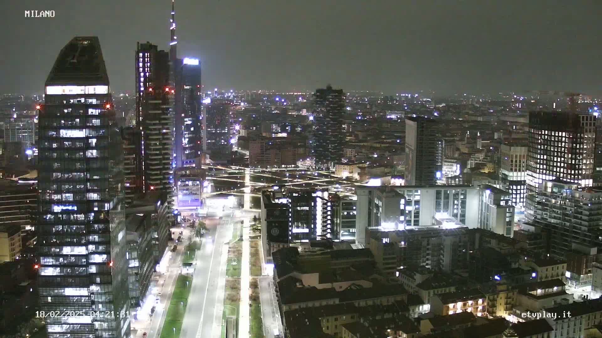 A nighttime aerial view of a city skyline showing numerous illuminated skyscrapers and buildings, with a wide, mostly empty street cutting through the scene under a dark, possibly overcast sky.