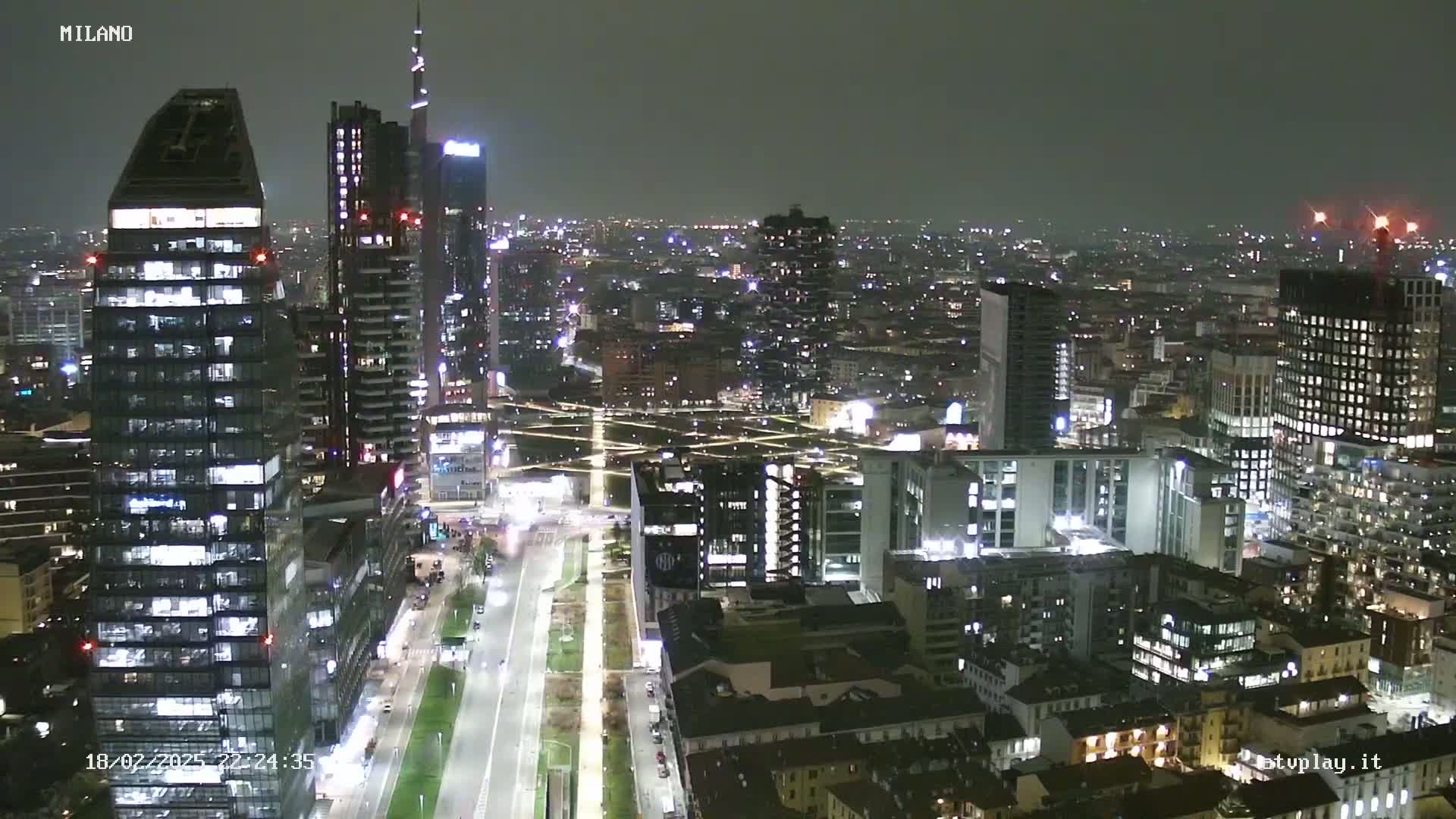 A nighttime aerial view of a city with many illuminated skyscrapers and buildings, under a dark, cloudy sky.