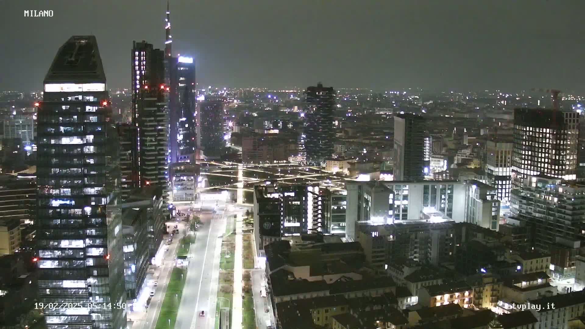 A nighttime aerial view of a city skyline with many illuminated skyscrapers and buildings, under a dark, clear sky.