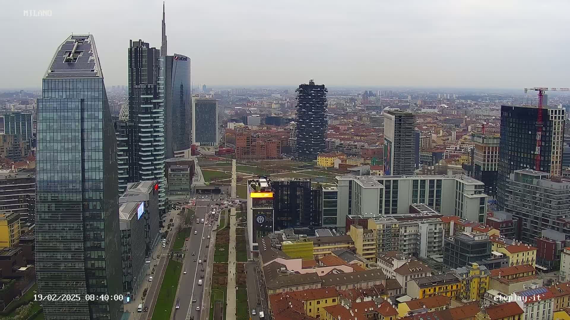 An aerial view of a city's skyline on an overcast day, showcasing numerous skyscrapers and high-rise buildings, with a wide road and park visible below.