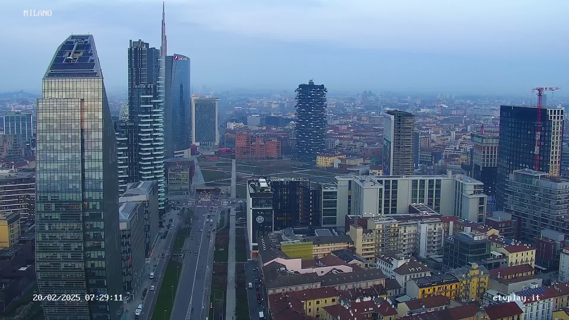 A hazy, overcast day reveals a cityscape of numerous modern skyscrapers and older buildings, with a long road running through the center.