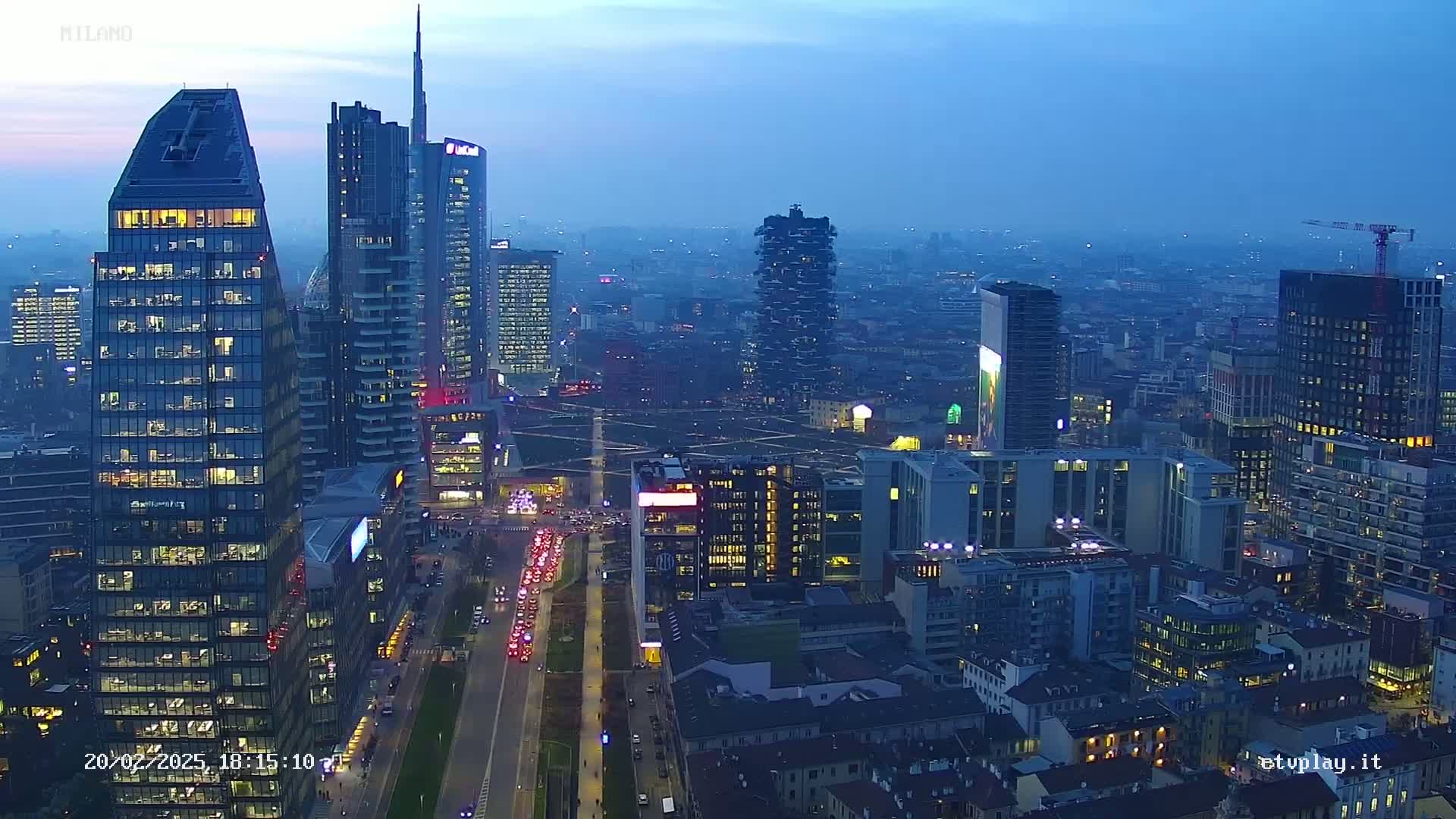 A twilight aerial view of a city skyline with numerous skyscrapers and city lights, showing a long road with traffic.
