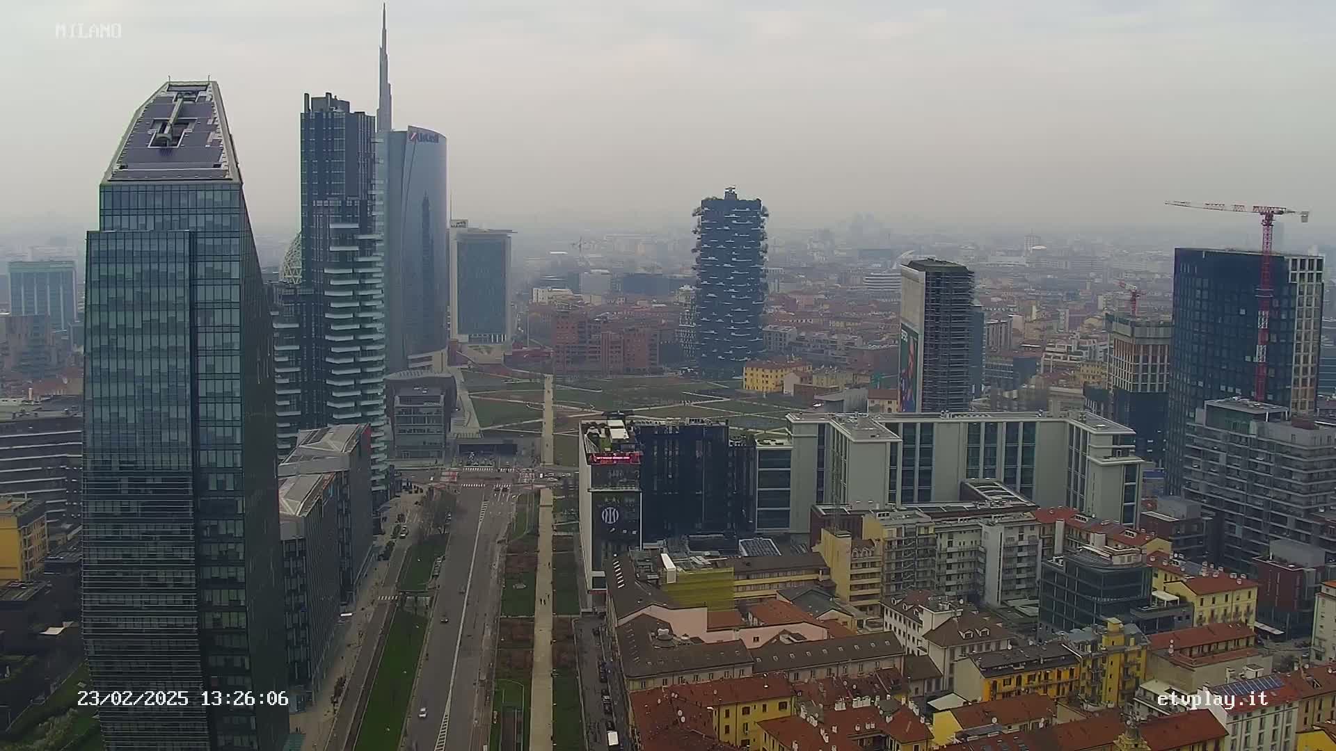 A hazy, overcast day reveals a dense cityscape of tall, modern skyscrapers and shorter, older buildings, with a wide street running through the center.