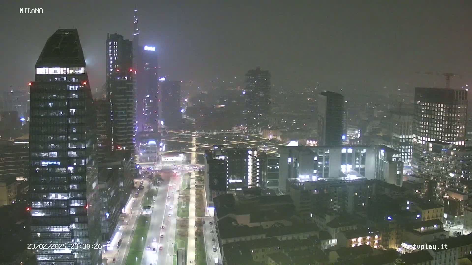 A nighttime view of a city skyline with many tall buildings, lit up under a hazy or foggy sky.