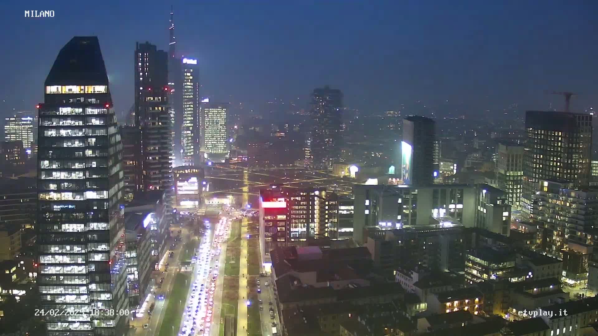 A nighttime aerial view of a city skyline showing numerous illuminated skyscrapers and a busy roadway with traffic under a dark, clear sky.