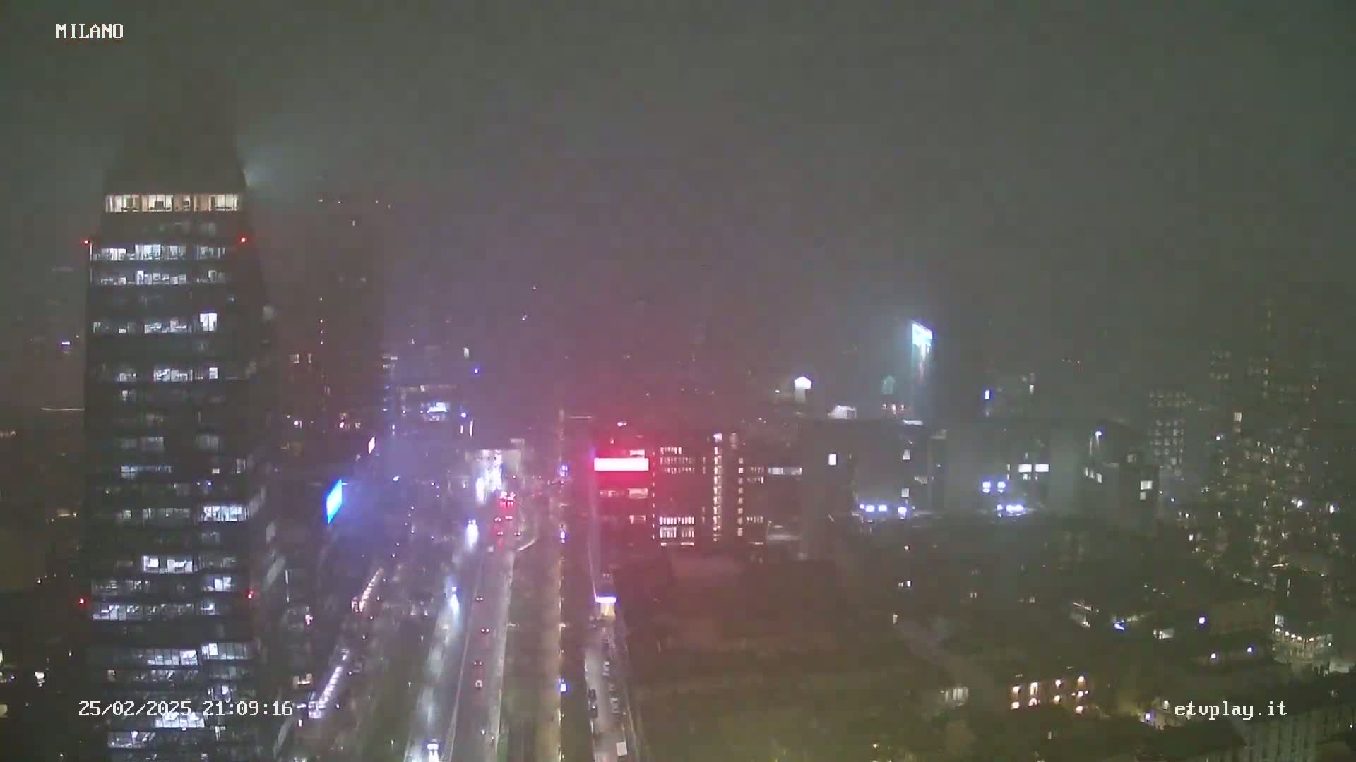 A nighttime, rain-blurred view of a city street with tall buildings and minimal traffic.