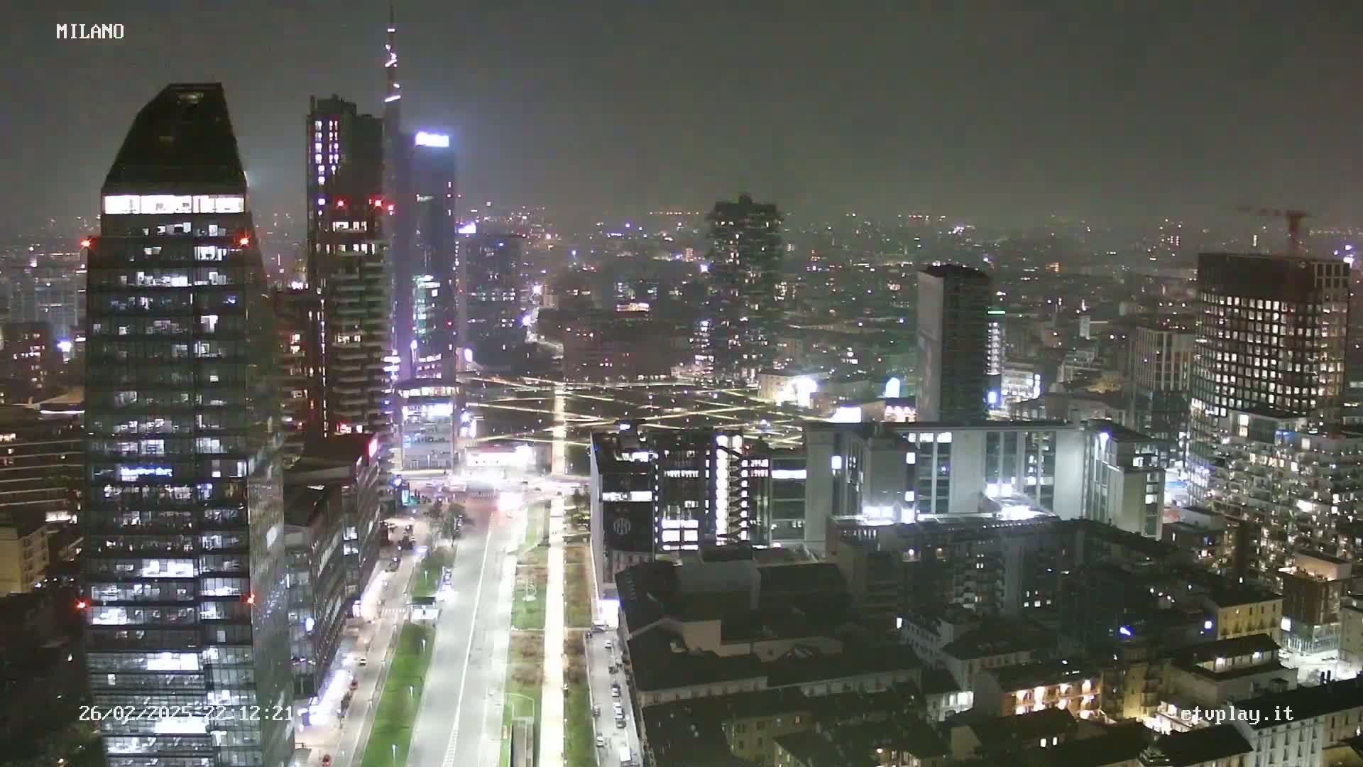A nighttime aerial view of a city skyline, showing numerous illuminated skyscrapers and streets under a hazy sky.