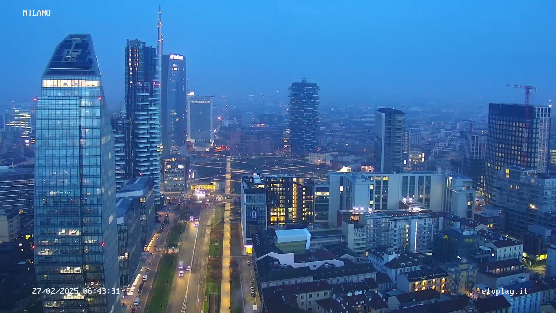 An aerial view of a city skyline at dusk, showing numerous skyscrapers and city buildings under a hazy, dark blue sky.