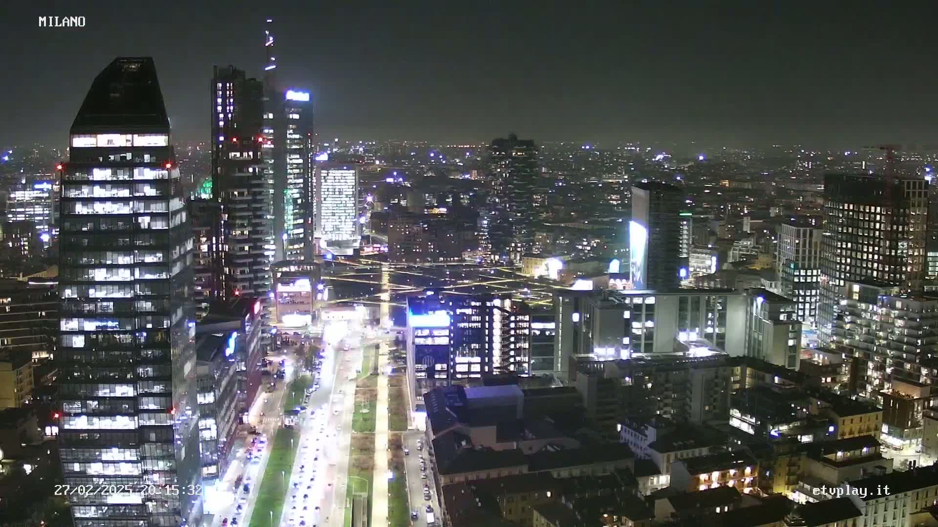 A nighttime aerial view of a city with many illuminated skyscrapers and streets showing vehicle traffic.