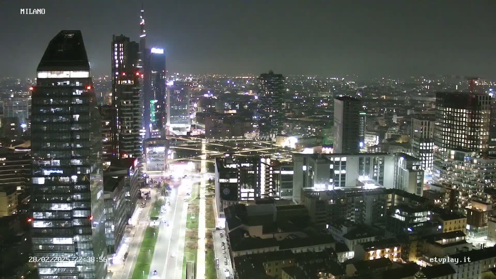 A nighttime aerial view of a city skyline showing numerous illuminated skyscrapers and buildings, with streets and open spaces visible below.