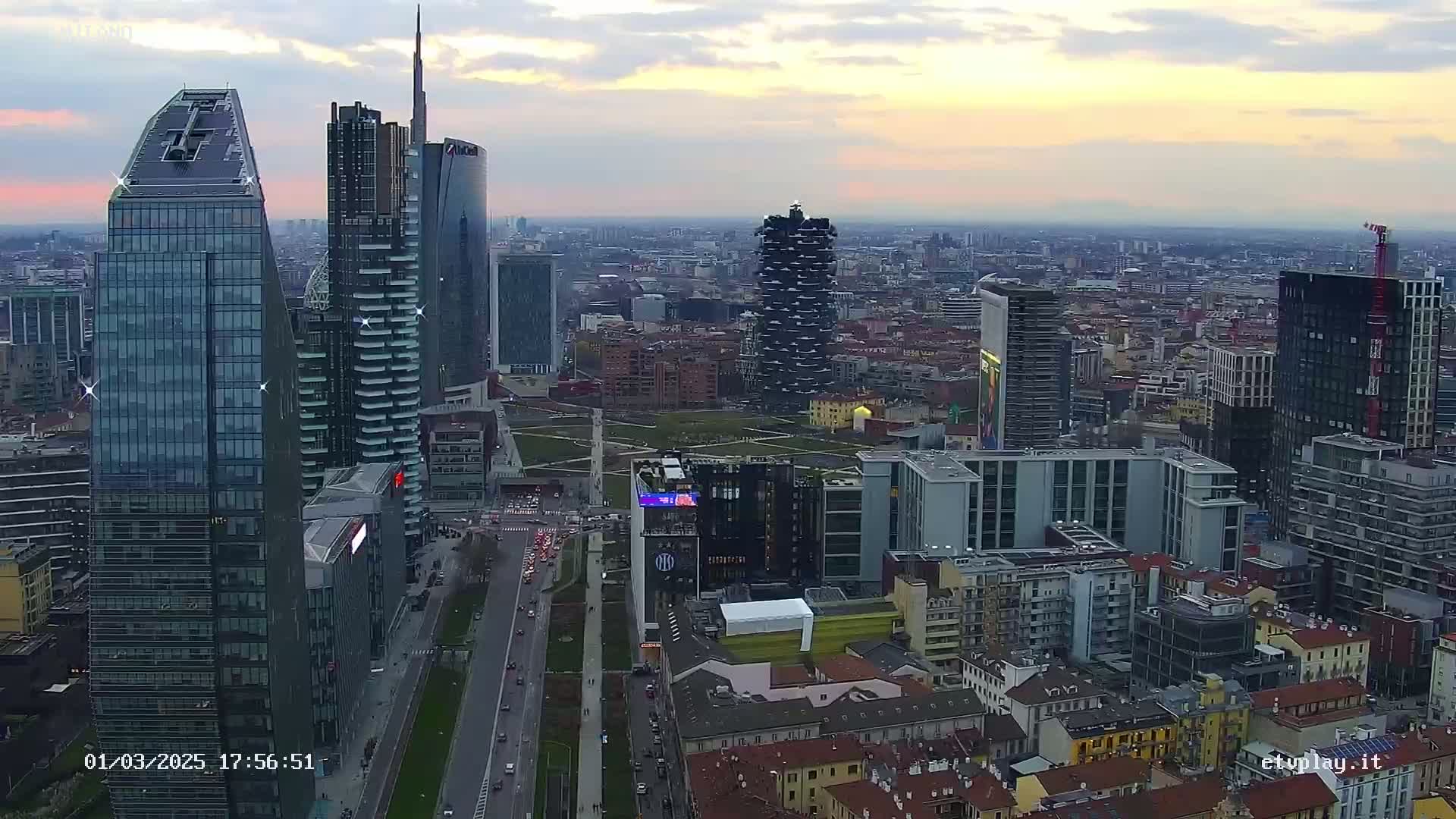 An aerial view of a dense cityscape at dusk, showing numerous skyscrapers and high-rise buildings under a cloudy sky.