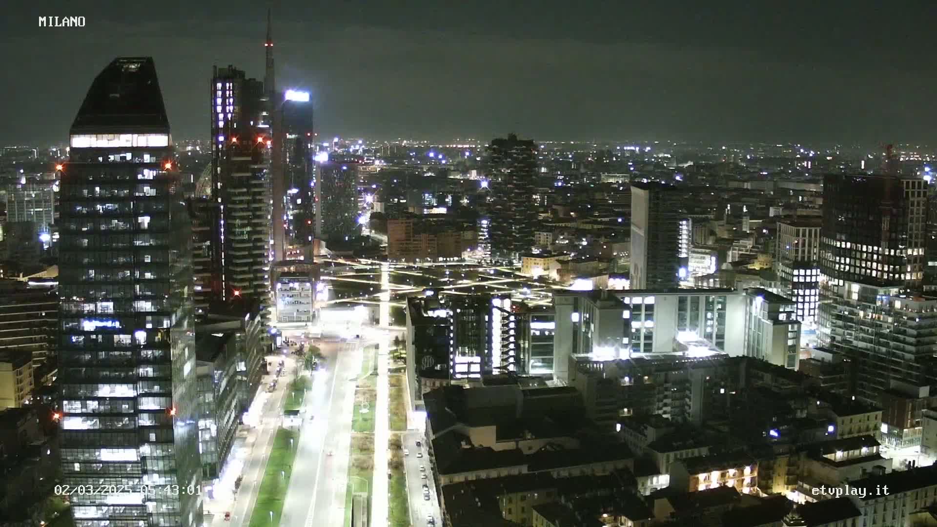 A nighttime aerial view of a city with numerous skyscrapers and brightly lit streets, under a clear sky.