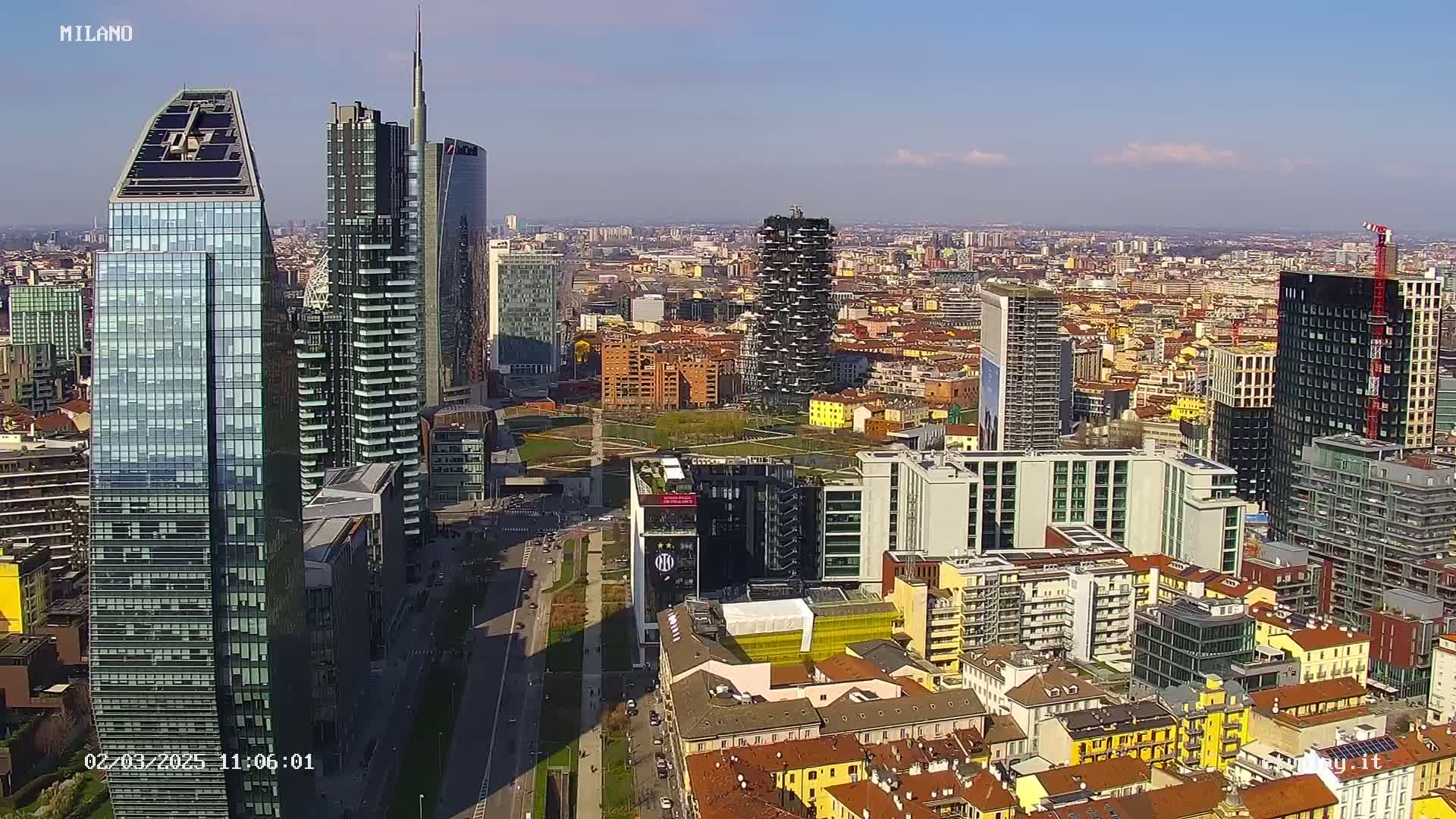 A high-angle view of a dense cityscape under a clear, sunny sky shows numerous skyscrapers and other buildings of varying heights and architectural styles.