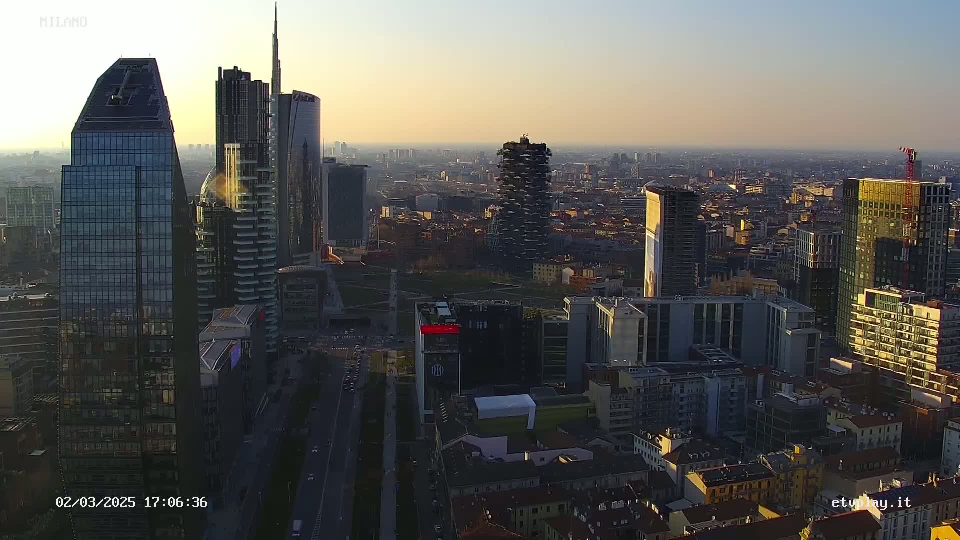 An aerial view of a dense cityscape at sunset, showing numerous skyscrapers and surrounding buildings under a clear sky.