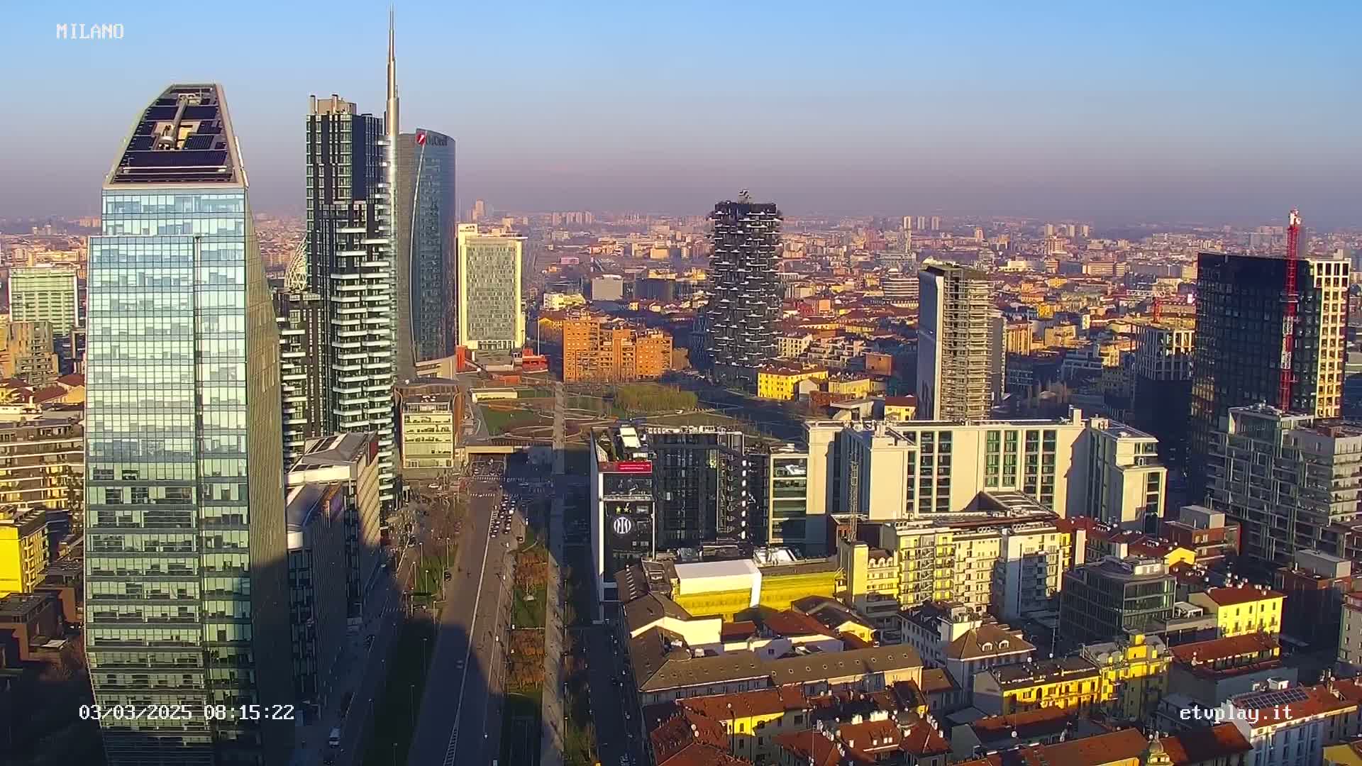 An aerial view of a dense city skyline on a clear, sunny day, showing many tall modern buildings and a mix of architectural styles.