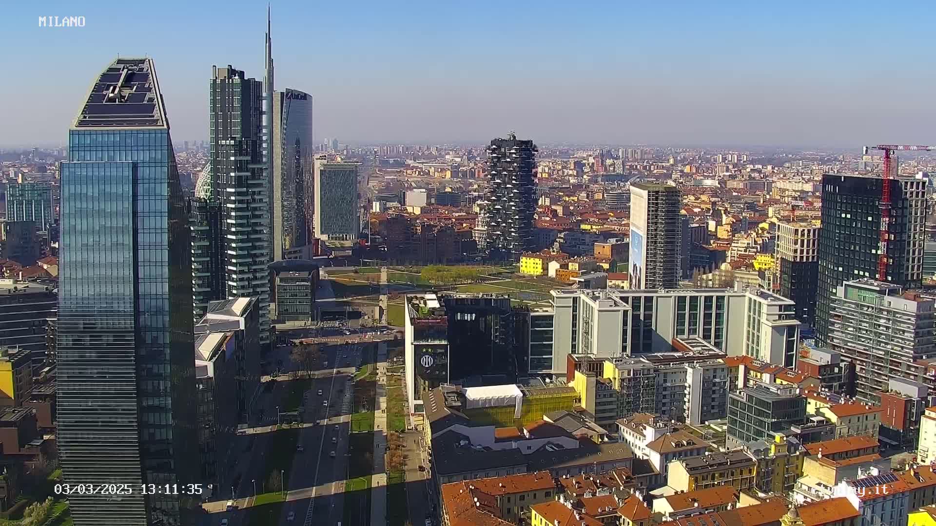 An aerial view of a city skyline on a sunny day, showing a cluster of modern skyscrapers and surrounding buildings.