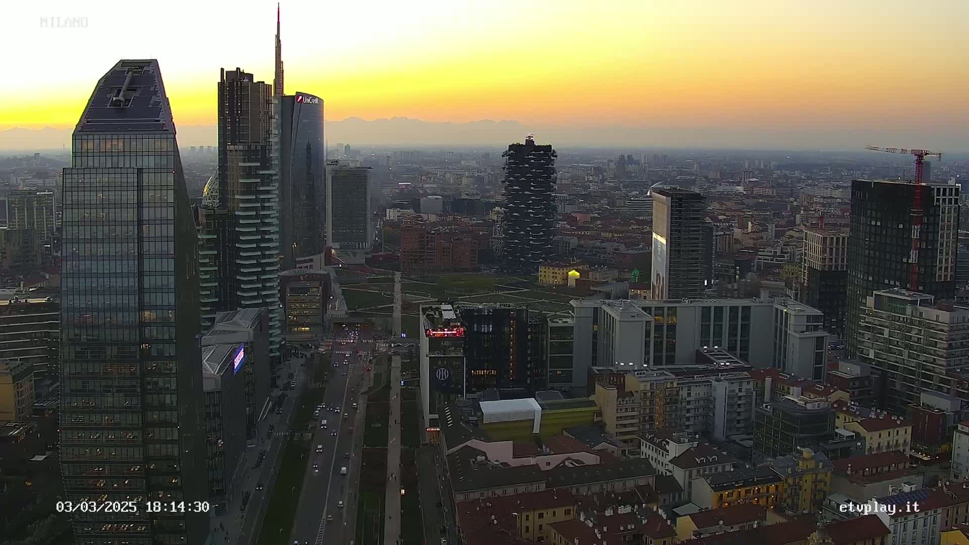 A high-angle, sunset view of a dense cityscape with numerous skyscrapers, featuring a mostly clear, yellow-orange sky.