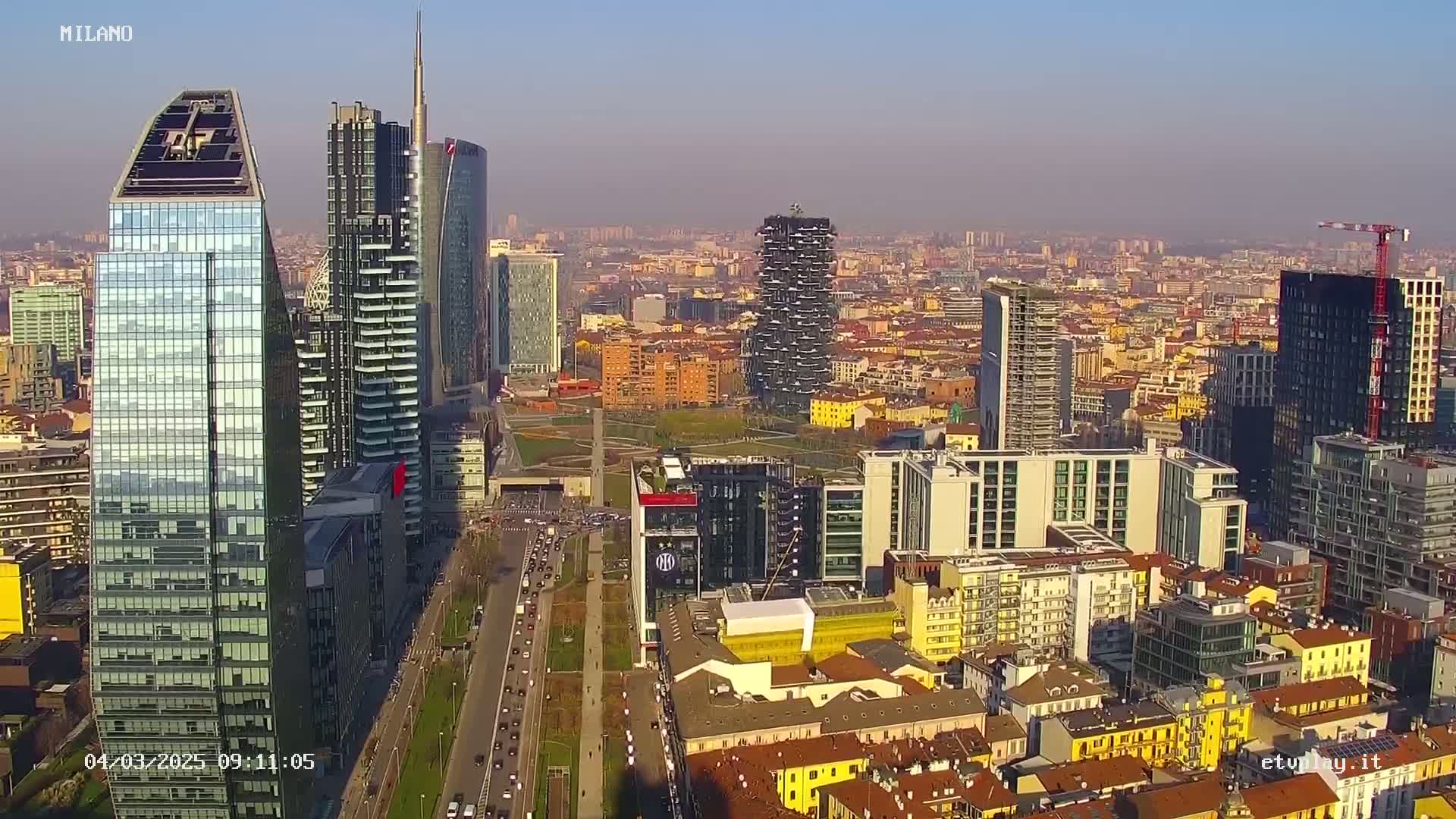 A high-angle, long shot of a city skyline on a clear, sunny day, showing numerous skyscrapers and other buildings, with a highway visible in the foreground.