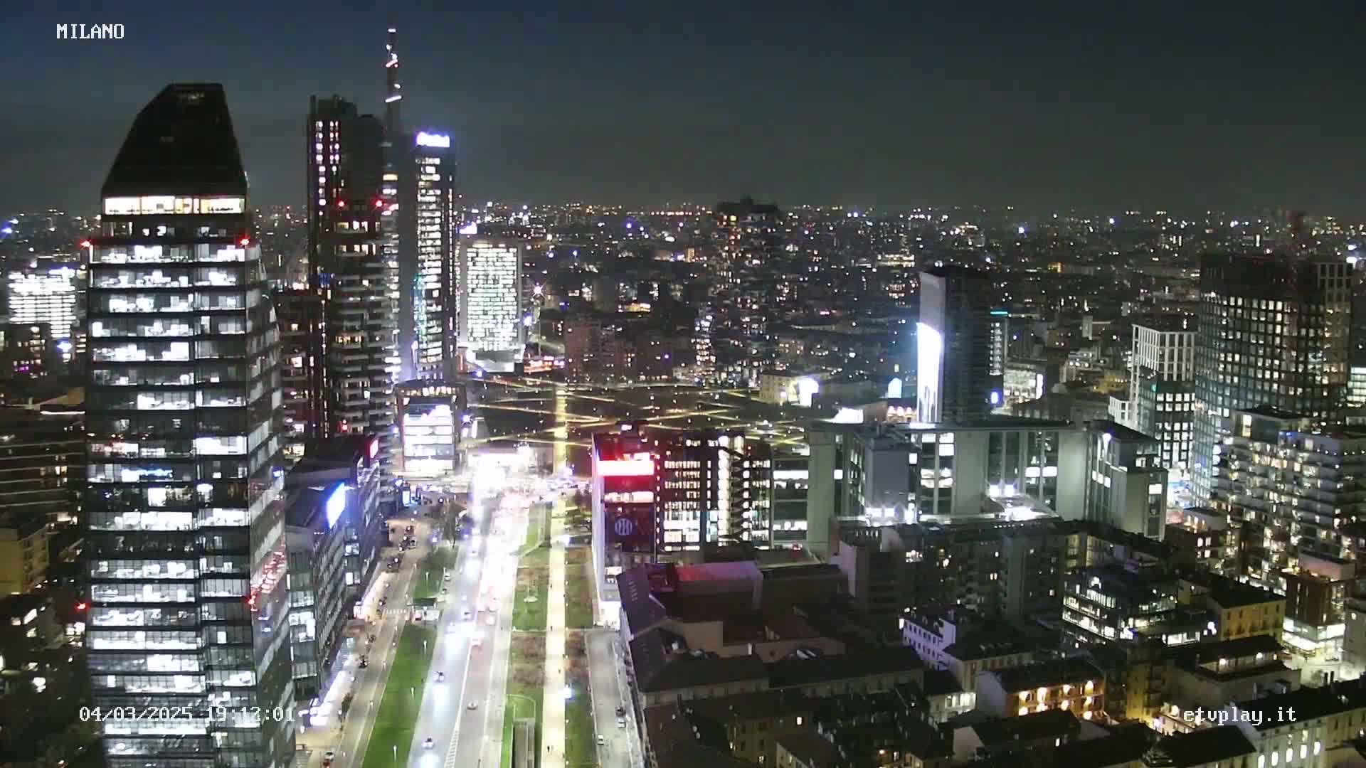 A nighttime, aerial view of a city's illuminated skyline, showing many skyscrapers and city streets with traffic.