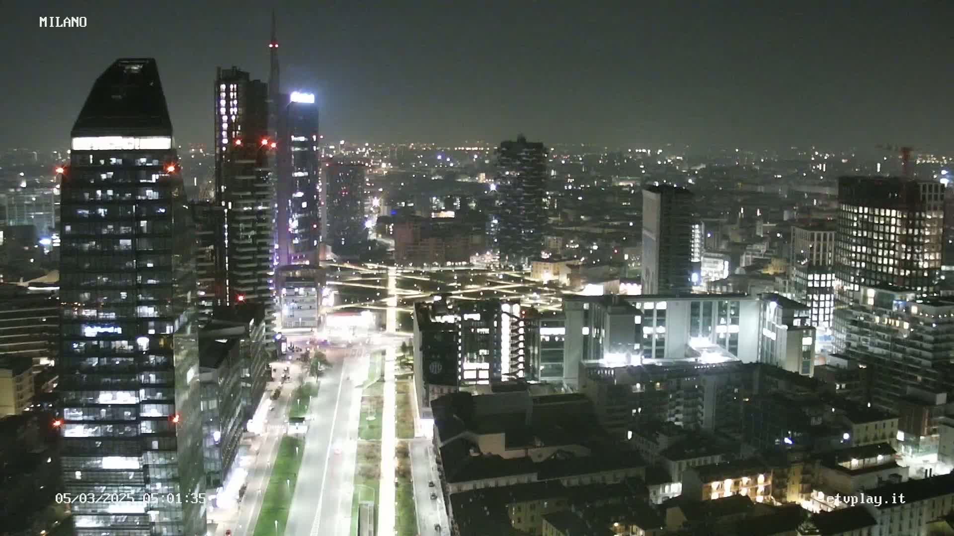 A nighttime aerial view of a city skyline, showing numerous illuminated skyscrapers and buildings, with a long, straight road cutting through a landscaped area.