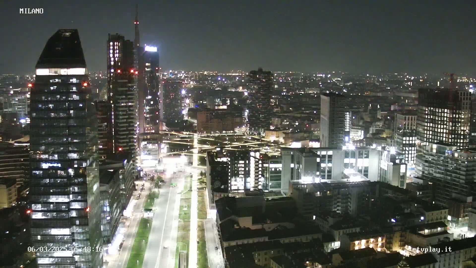 A nighttime aerial view of a city with many illuminated skyscrapers and buildings, showing a well-lit road cutting through a green space.