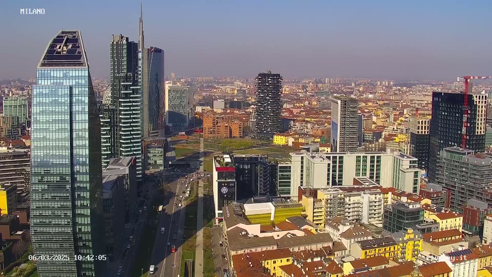 A high-angle, long shot of a city skyline on a clear, sunny day, showing many skyscrapers and other buildings, with a wide street and park visible below.