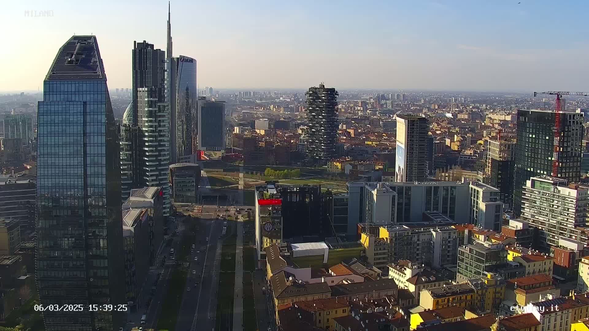 A high-angle, long shot shows a city skyline of many modern skyscrapers and shorter buildings under a clear, sunny sky.