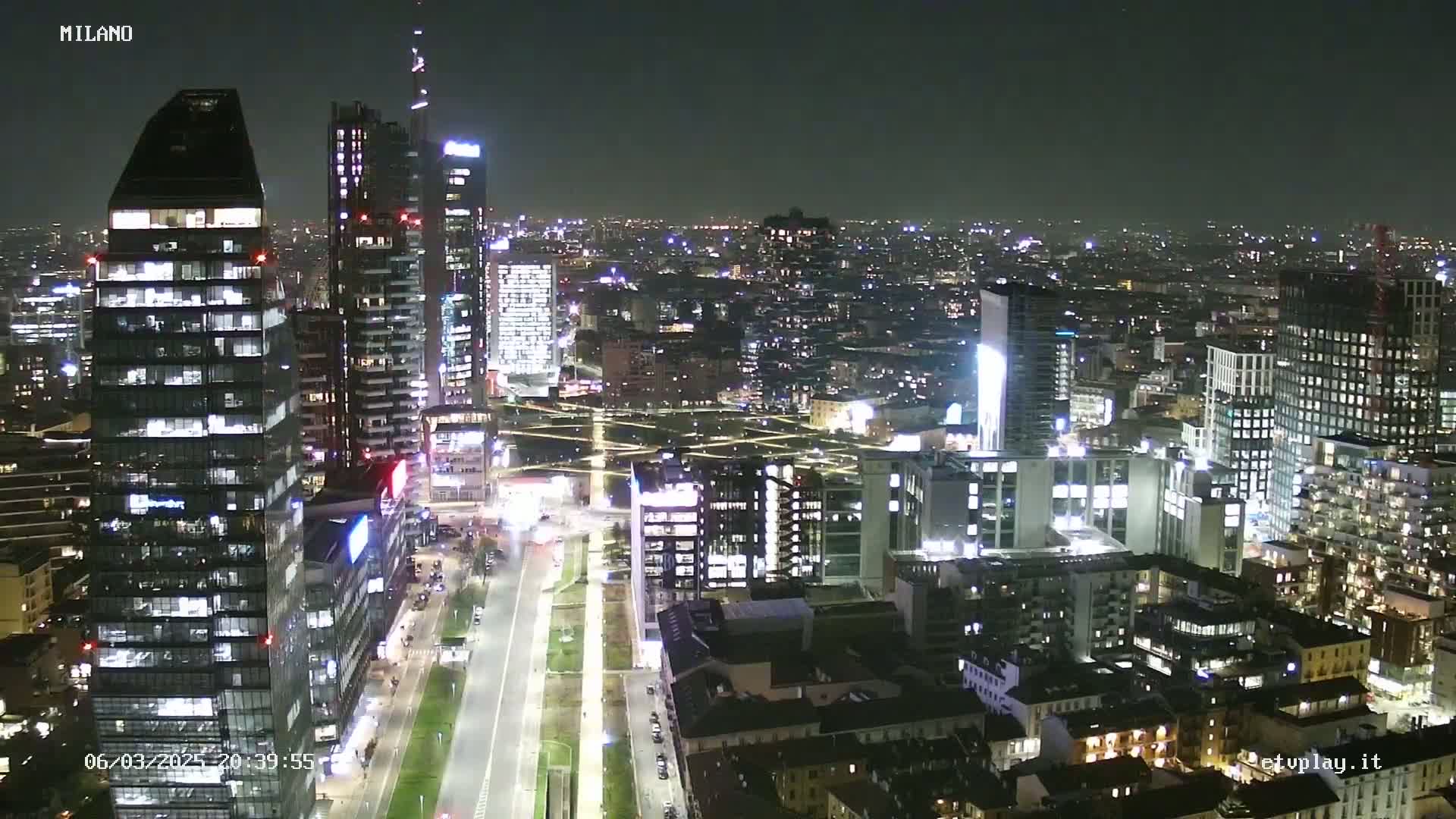 A nighttime aerial view of a city, showing many illuminated skyscrapers and buildings stretching into the distance under a clear, dark sky.
