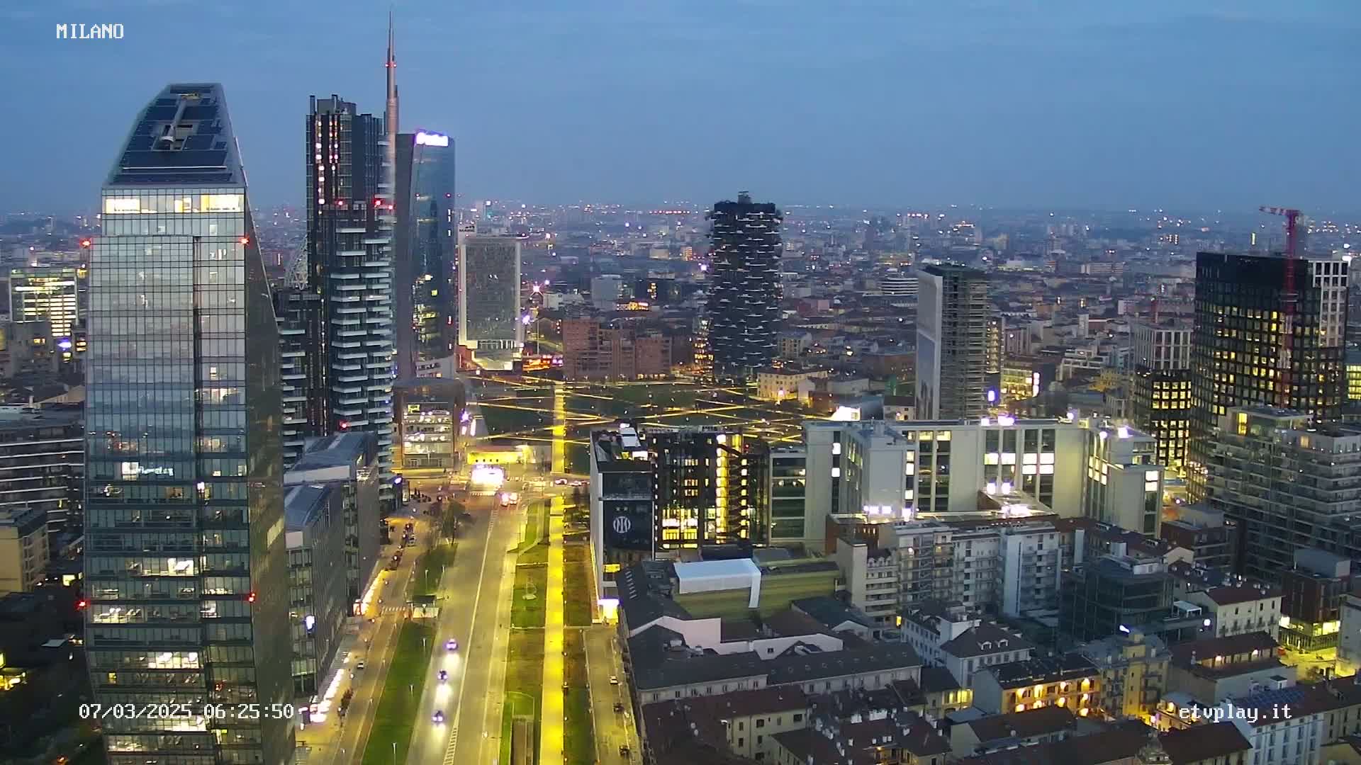 A nighttime aerial view of a city with numerous skyscrapers and high-rise buildings, illuminated under a twilight sky.