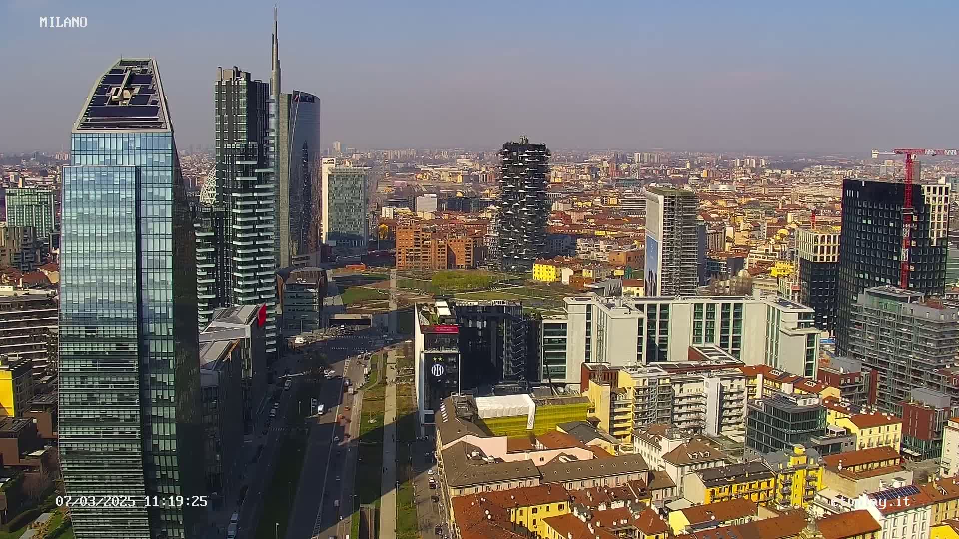 An aerial view of a city skyline on a sunny day, showing numerous skyscrapers and surrounding buildings.