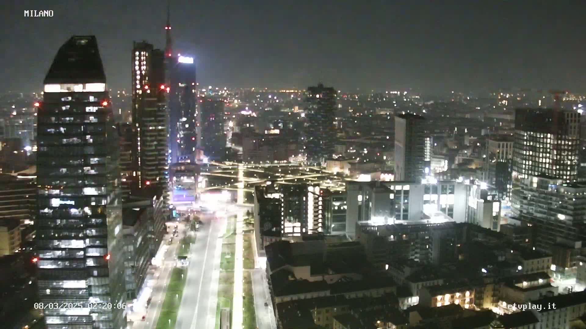 A nighttime aerial view of a city skyline showing numerous illuminated skyscrapers and buildings, with a long, brightly lit road cutting through the scene.