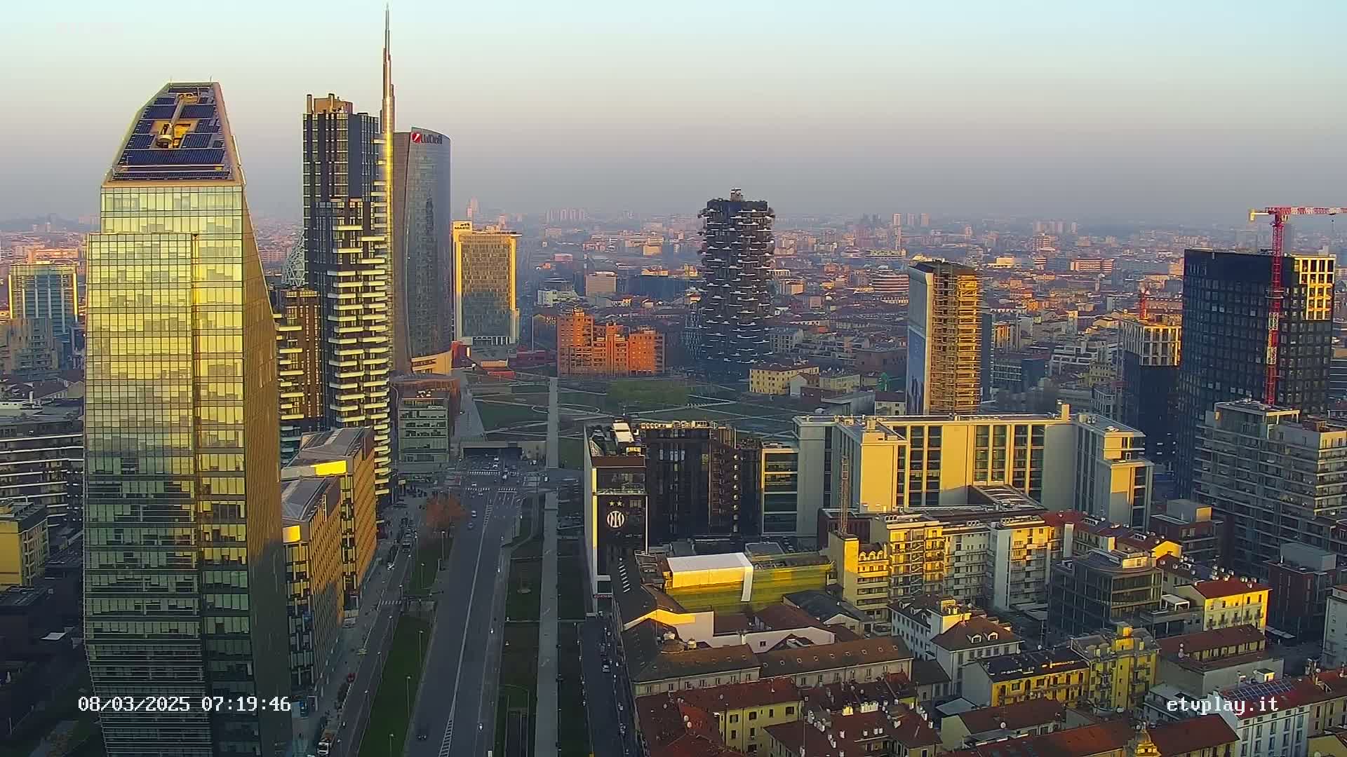 An aerial view of a city skyline at sunset, showing numerous skyscrapers and buildings of varying heights, with hazy atmospheric conditions.