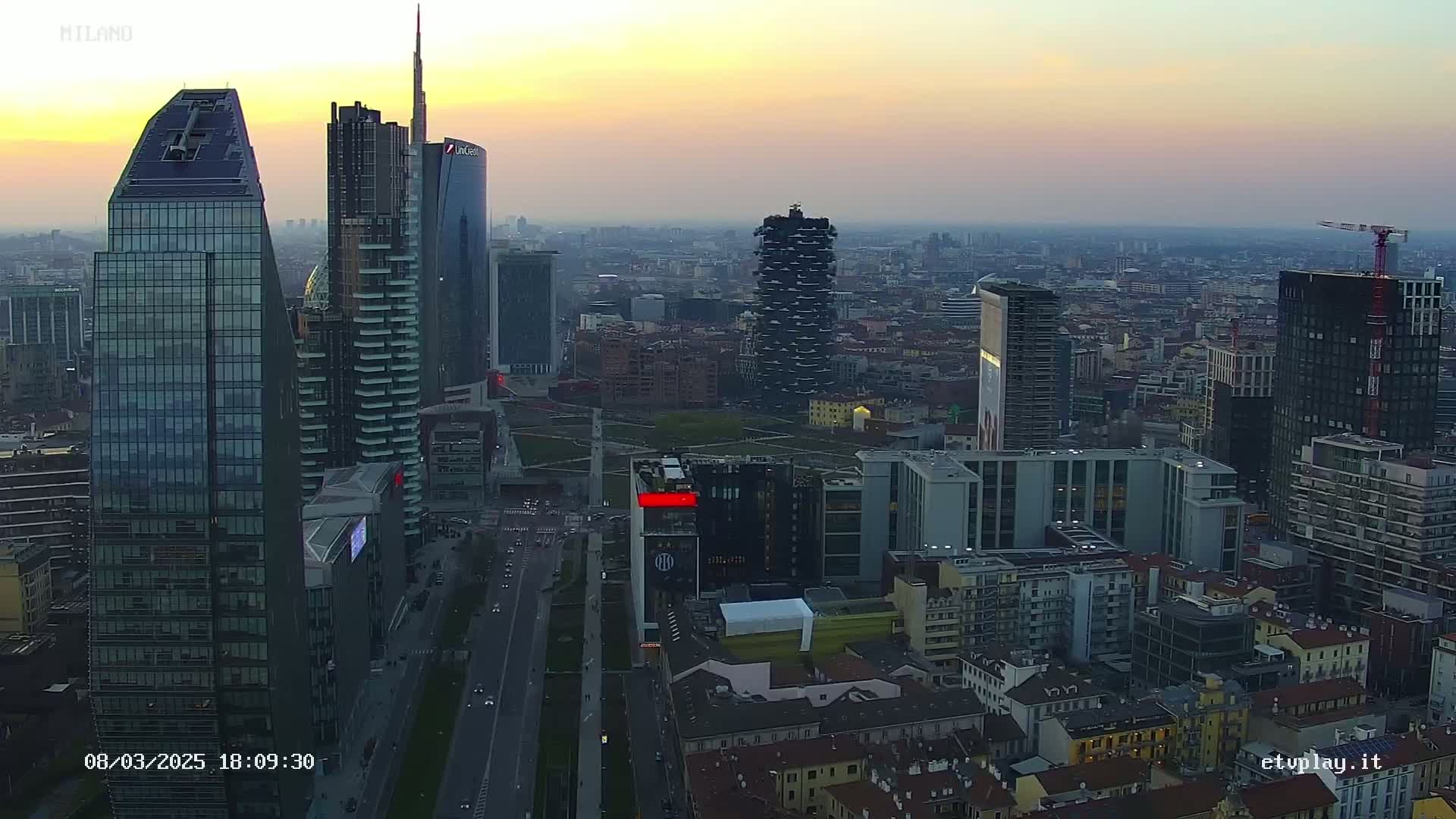 An aerial view of a dense cityscape at sunset, showing numerous skyscrapers and surrounding buildings under a hazy sky.